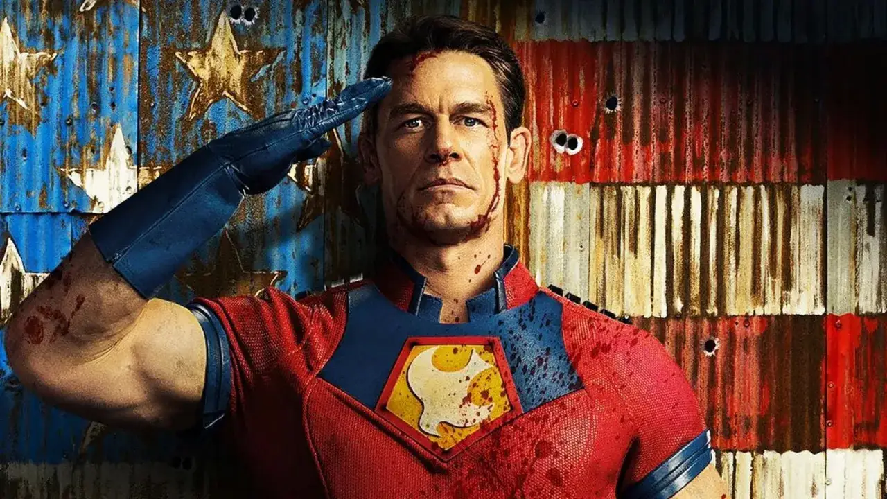 John Cena w kostiumie Peacemakera, z krwią na twarzy i ubraniu, salutuje na tle zniszczonej flagi USA. Peacemaker obsada w akcji.