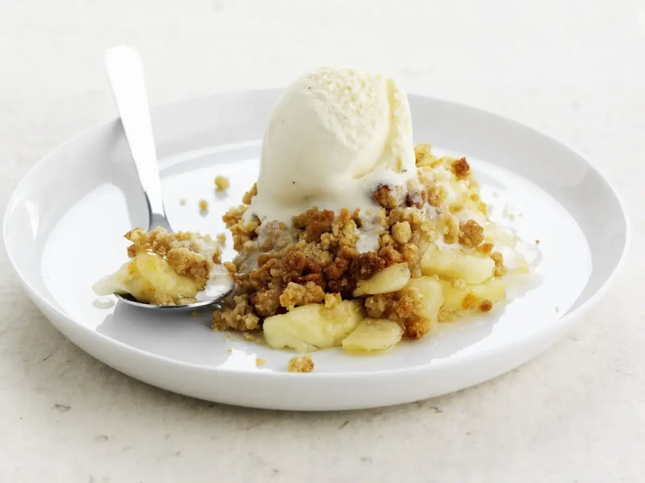 Ein L&ouml;ffel voller warmem Apple Crumble mit einer Kugel Vanilleeis, das langsam schmilzt.