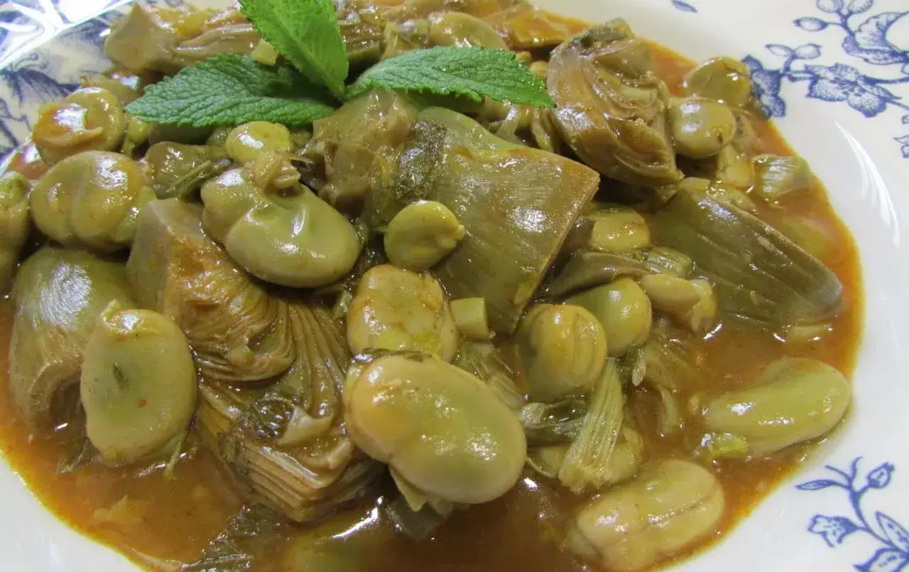 Plato de habas tiernas y trozos de alcachofa en una salsa dorada, decorado con hojas de menta fresca.