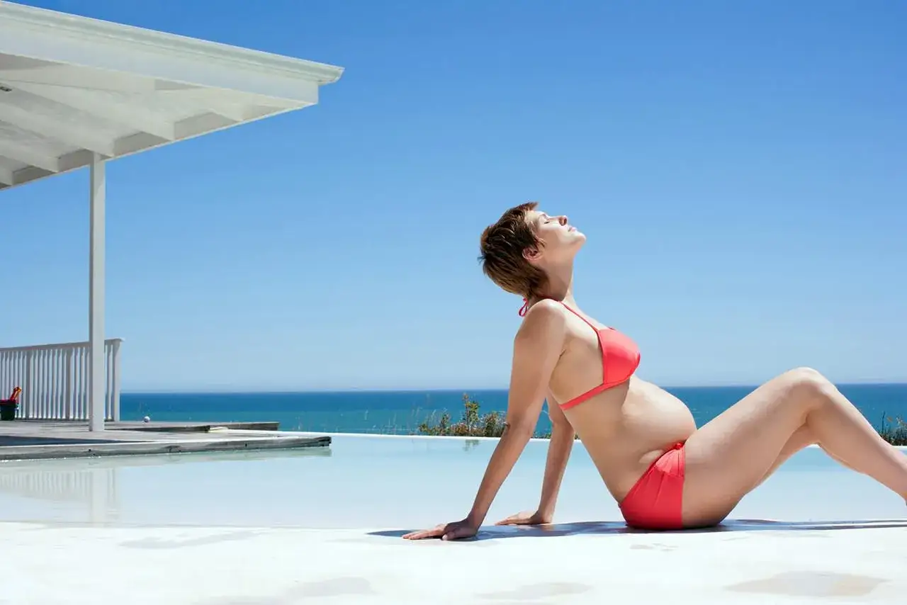Kobieta w bikini cieszy się opalaniem w ciąży przy basenie z widokiem na ocean.