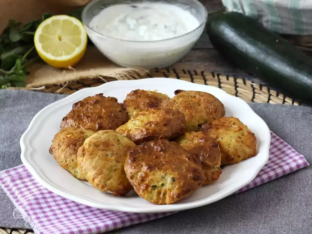 Gyllene, friterad zucchini-kakor på en vit tallrik, serverade med en klyfta citron och en skål med krämig dipp.