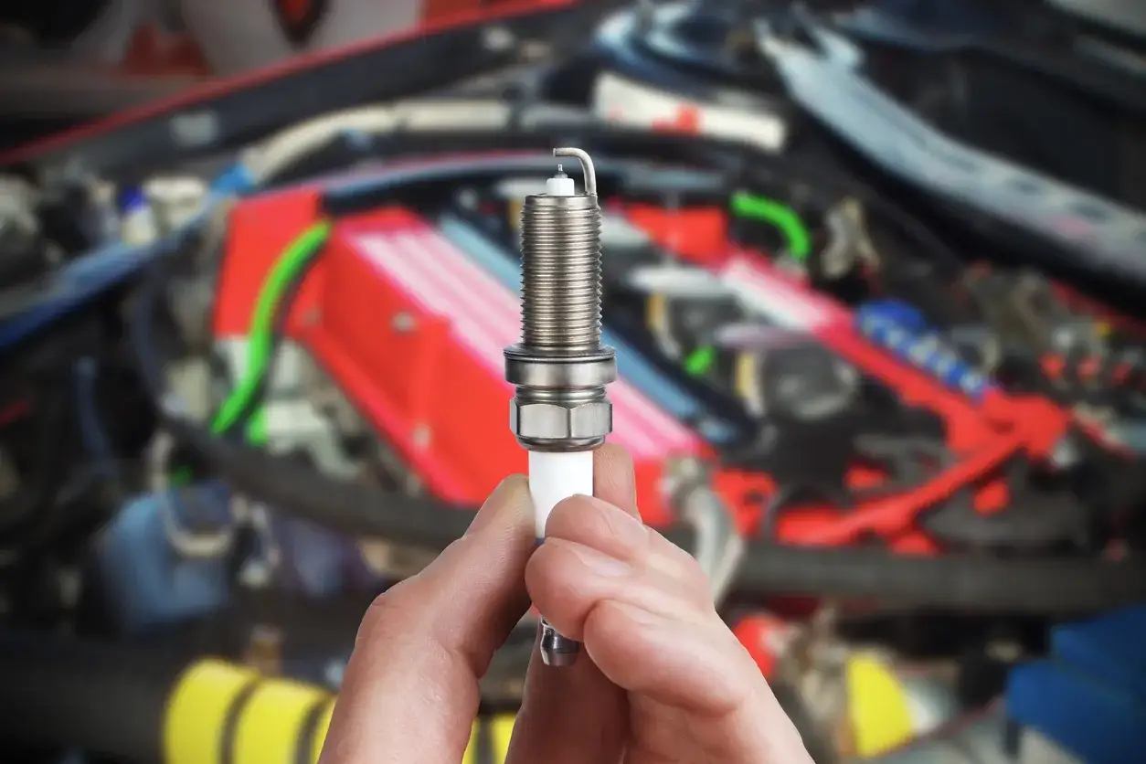 Une bougie d'allumage tenue en main, possible cause d'une voiture qui broute au d&eacute;marrage. Le moteur est flou en arri&egrave;re-plan.