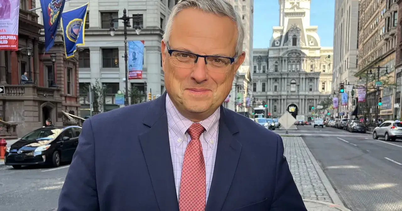 Marcin Wrona w garniturze i krawacie, na tle ulicy z budynkami i samochodami. Może to jego żona zrobiła to zdjęcie.