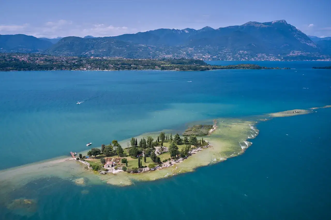 Un'incantevole isola nel Lago di Garda, con case, alberi e un molo, circondata da acque turchesi e montagne sullo sfondo.