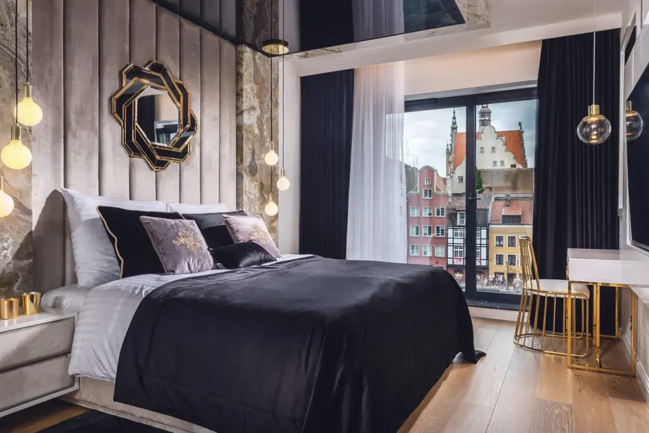 Elegancka sypialnia glamour z widokiem na urokliwe kamienice. Miękkie poduszki, luksusowa pościel i złote akcenty tworzą przytulną atmosferę.