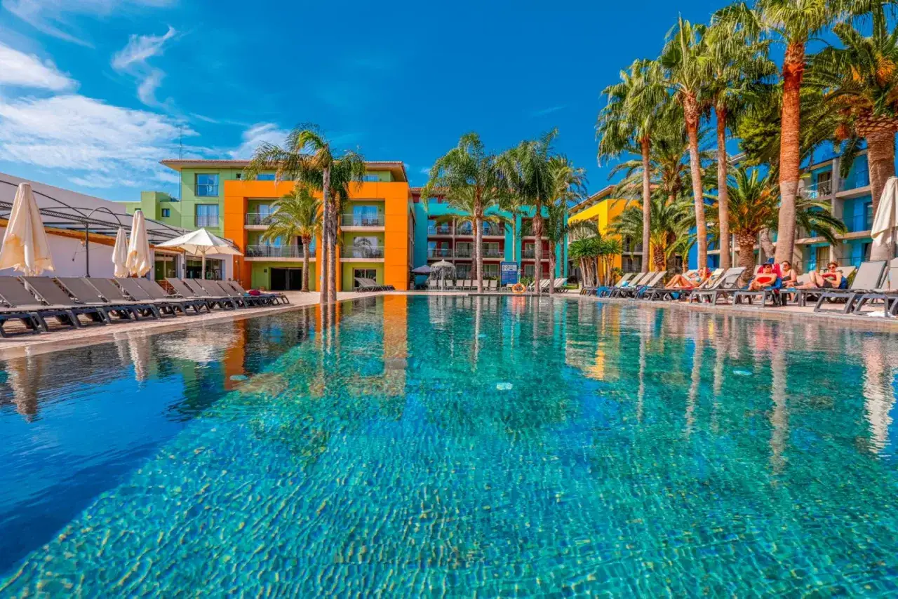Piscina scintillante e palme rigogliose al **hotel Occidental Menorca**, con edifici colorati sullo sfondo e persone che si rilassano sui lettini.