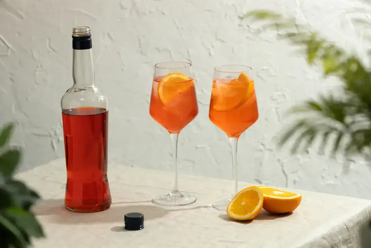Dwa kieliszki orzeźwiającego aperol spritz z plasterkami pomarańczy, obok butelka likieru i świeże owoce.