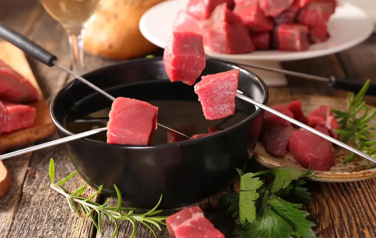 Des cubes de b&oelig;uf sur des brochettes pr&ecirc;ts &agrave; &ecirc;tre plong&eacute;s dans l'huile chaude pour une fondue bourguignonne. Un verre de vin blanc est visible en arri&egrave;re-plan.