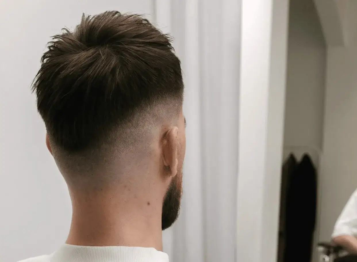 Cabello oscuro con un corte moderno, un **mid fade en pico** que se difumina suavemente hacia la nuca.