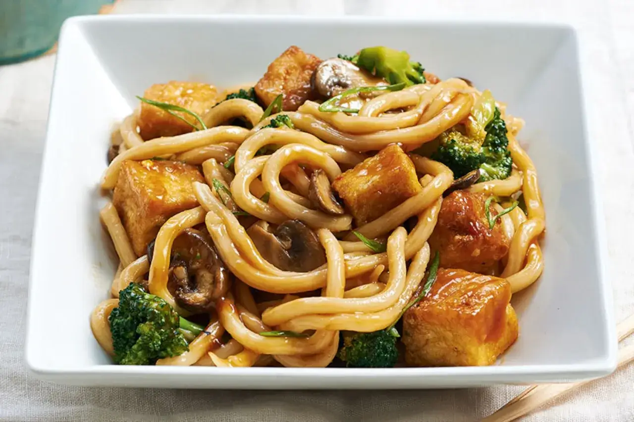 Herzhafte udon nudeln mit tofu, Brokkoli und Pilzen in einer köstlichen Sauce, serviert in einer weißen Schüssel.