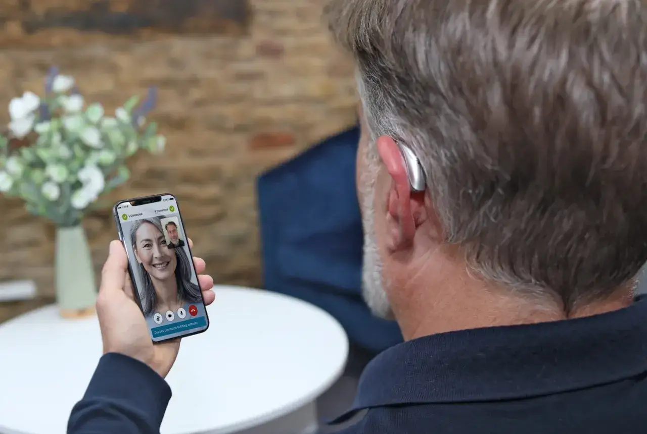 Mężczyzna z aparatem słuchowym rozmawia przez wideorozmowę na telefonie. Aplikacja na telefon pozwala na zdalne dopasowanie aparatów.