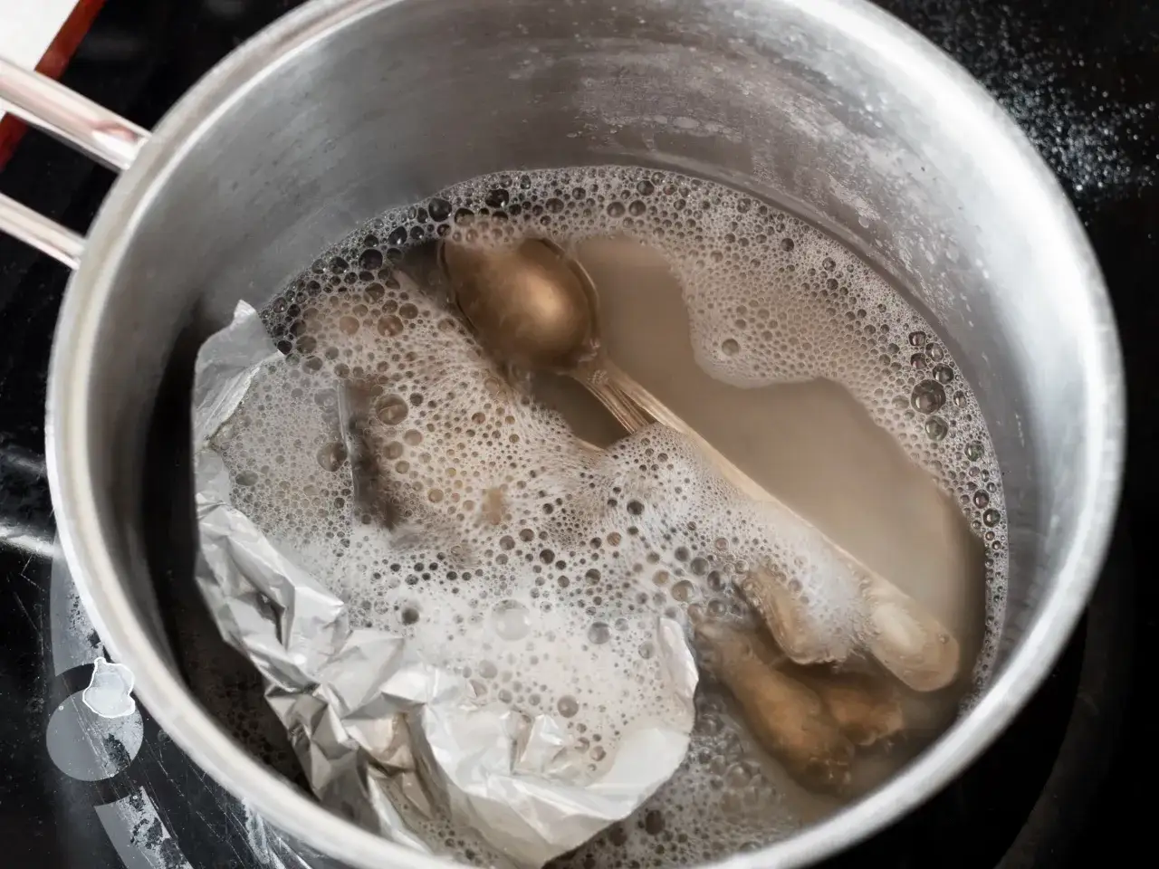 Czyszczenie sztućc&oacute;w sodą w garnku z folią aluminiową. Woda z mydłem tworzy pianę, a sztućce zaczynają lśnić.