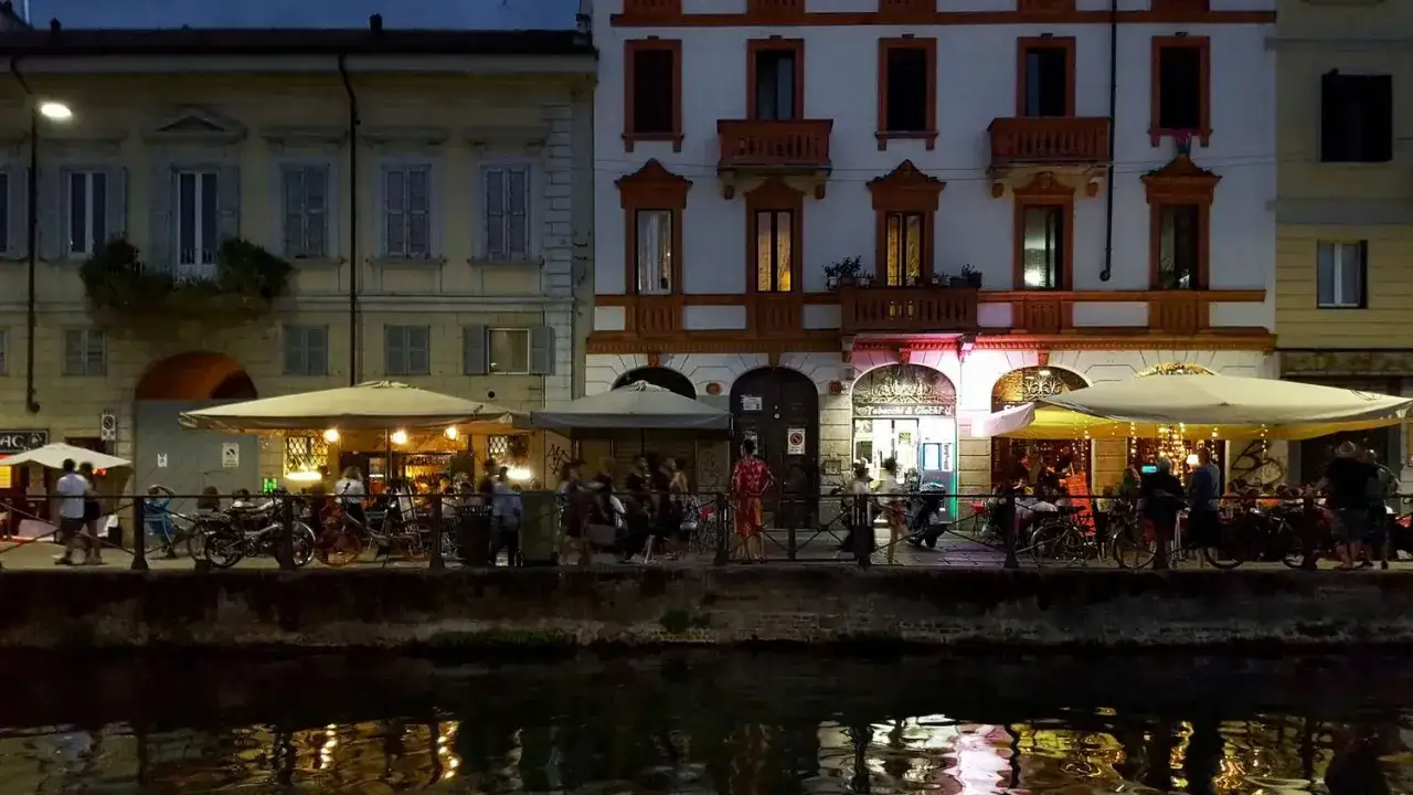 Serata sui Navigli, tra i locali trendy di Milano. Gente seduta ai tavolini all'aperto, luci soffuse e riflessi sull'acqua.