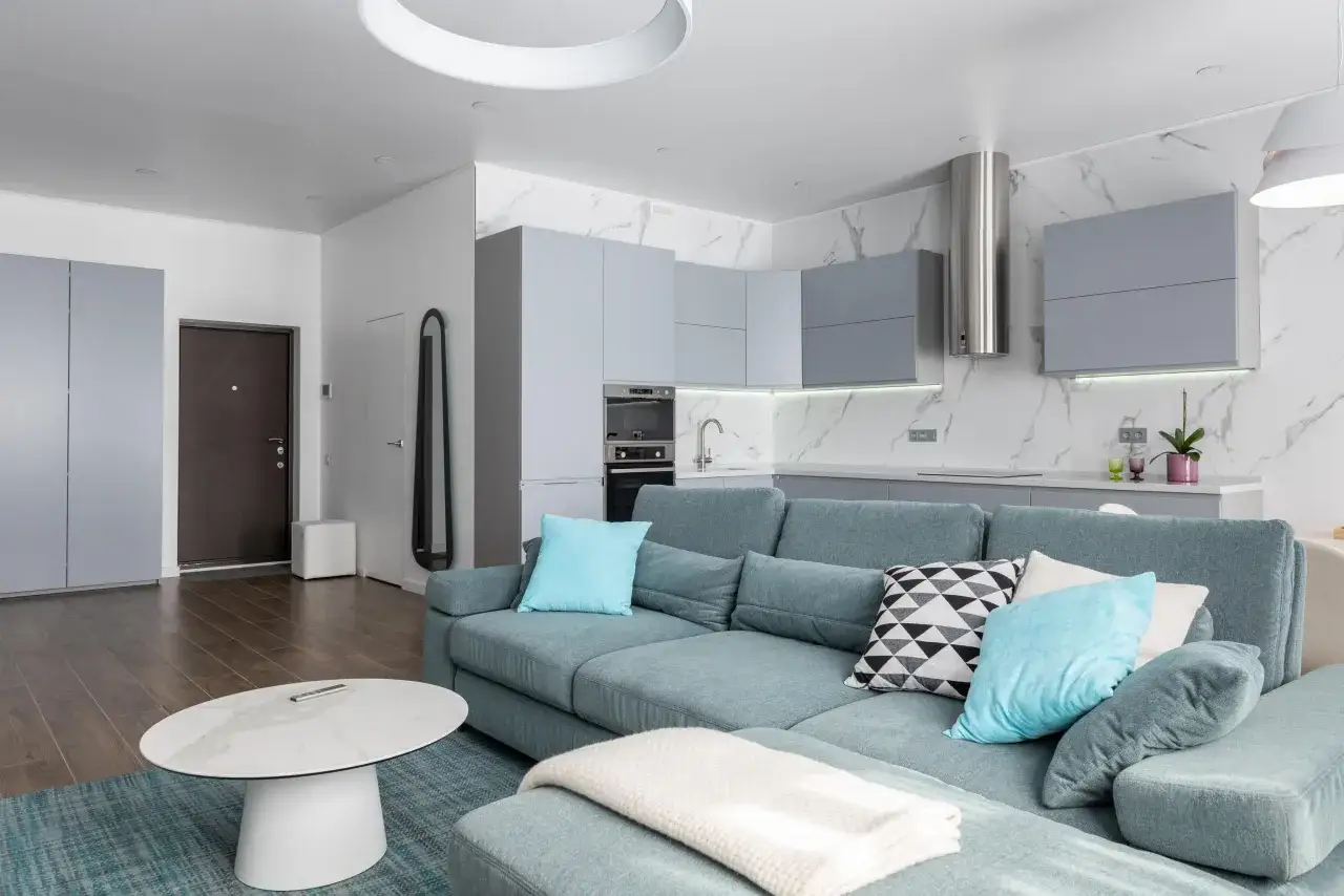 Espaciosa cocina sal&oacute;n comedor de 30 m2 con sof&aacute; azul, mesa de centro ovalada y cocina moderna con armarios grises.
