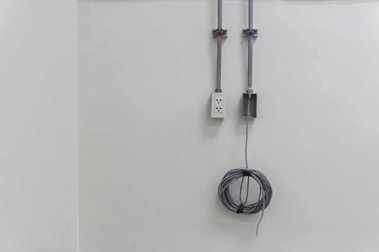 Dwa pionowe szare kable przymocowane do jasnej ściany. Obok gniazdko elektryczne i zwinięty kabel. Pokazuje, jak przymocować kabel do ściany.
