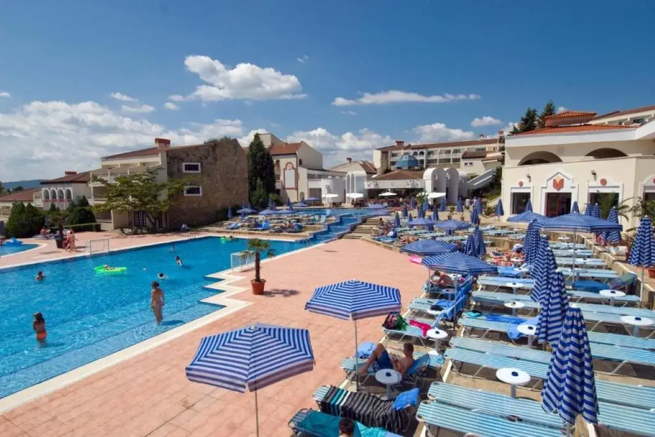 Basen hotelowy z leżakami i parasolami, w tle budynki hotelowe. Duni Royal Pelican zaprasza do relaksu.