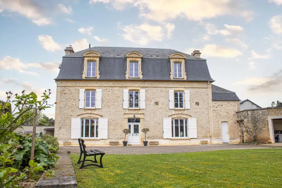 Belle maison en pierre avec une **installation de volets battants** blancs, un jardin verdoyant et un ciel nuageux.