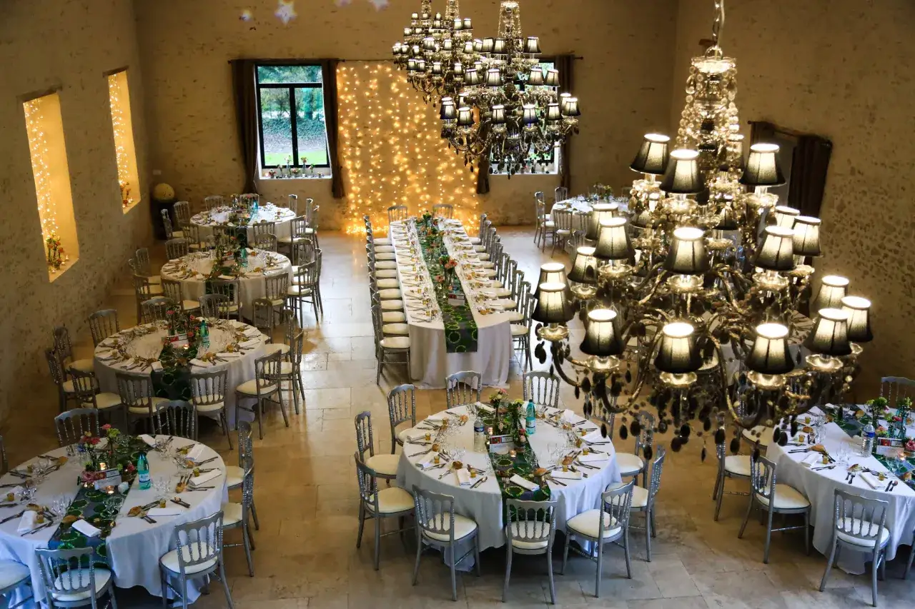 Plan de salle mariage : tables rondes dress&eacute;es, chaises argent&eacute;es, lustres imposants, guirlandes lumineuses dans une grange am&eacute;nag&eacute;e.