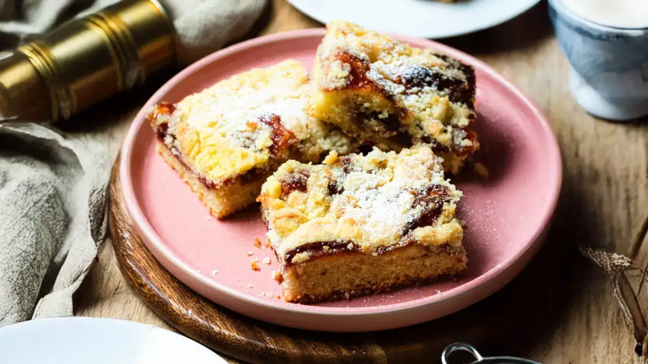 Saftiger Streuselkuchen mit Marmelade, bestäubt mit Puderzucker, auf einem rosa Teller.