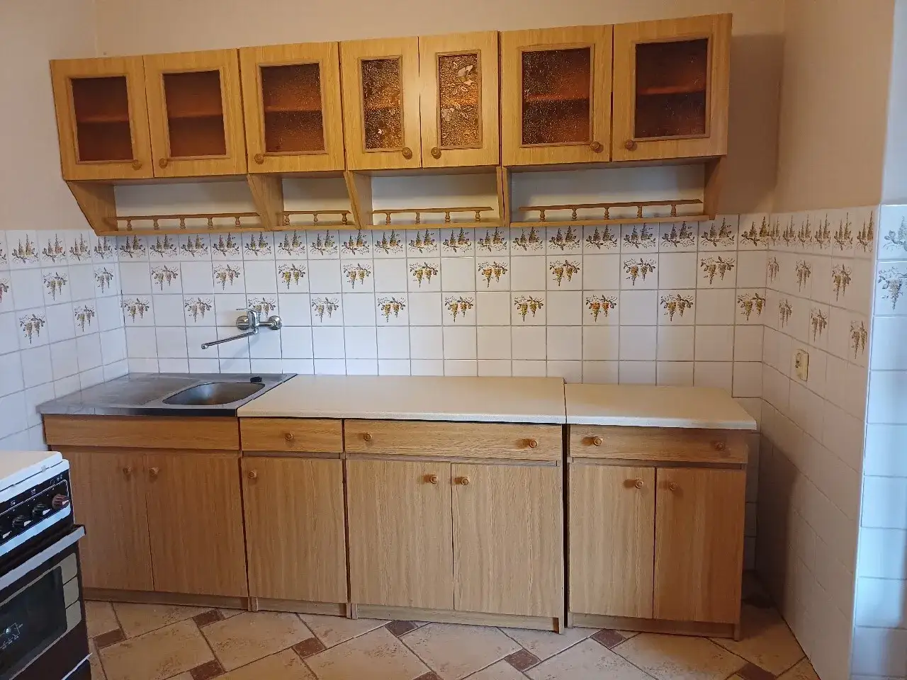 Jasne, oto opis:
Używane meble kuchenne z jasnego drewna, z przeszklonymi frontami szafek wiszących i ceramicznymi płytkami z motywem winogron.