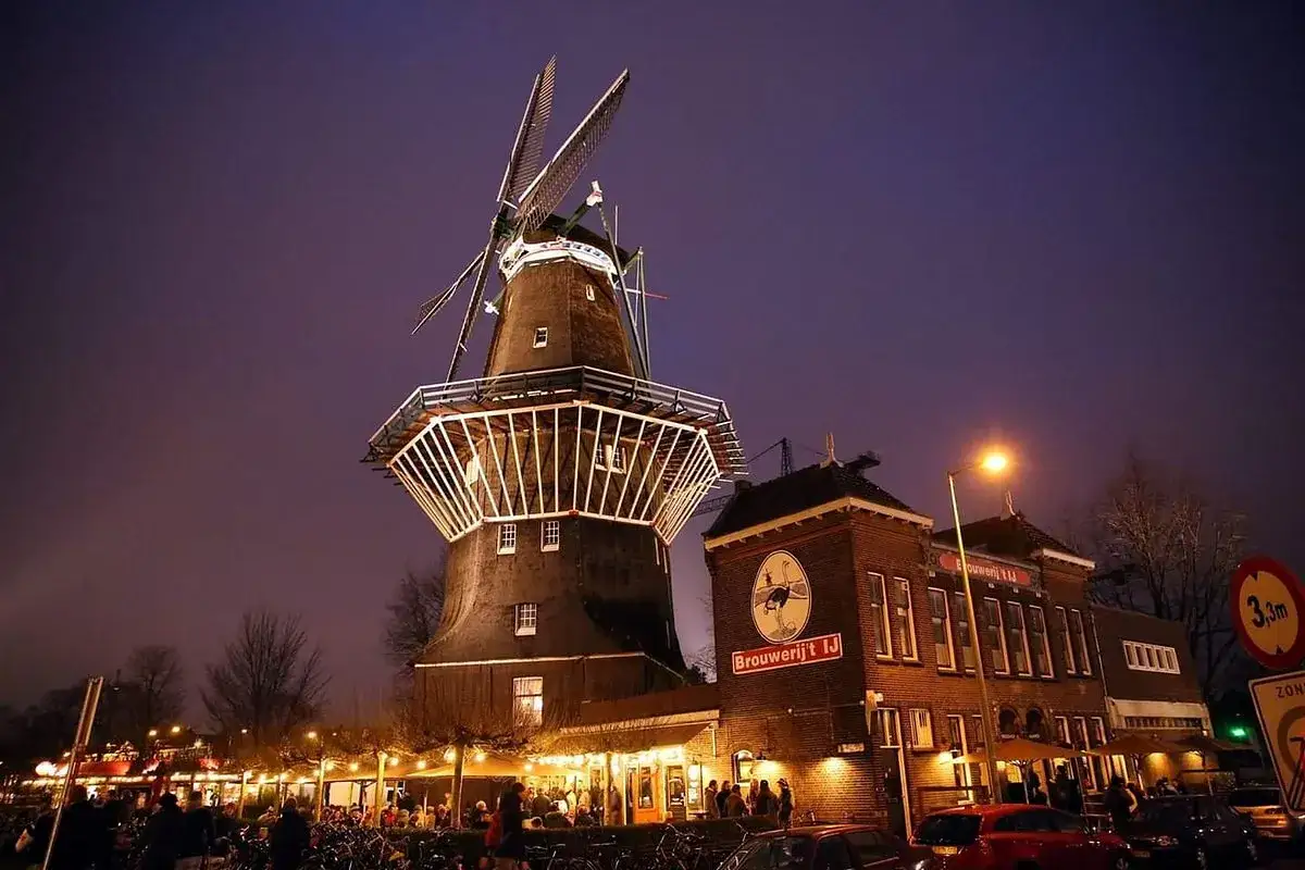 Mulino a vento illuminato di notte accanto alla Brouwerij 't IJ, con persone e biciclette all'esterno.