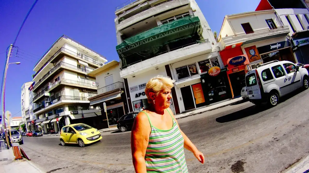 Donna cammina per strada, con edifici e negozi sullo sfondo. Una piccola auto gialla, un furgone bianco e altre auto sono visibili. L'immagine &egrave; stata catturata con una GoPro **Ace Pro 2**.