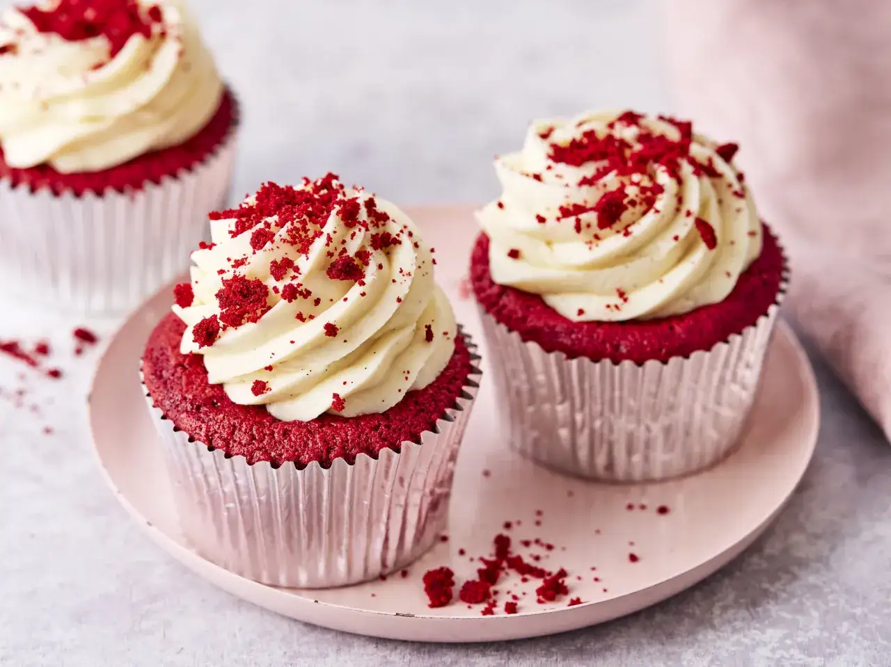 Zwei k&ouml;stliche red velvet Muffins mit cremiger Glasur und roten Kr&uuml;meln auf einem rosa Teller.