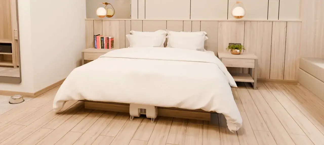 A white fan is placed under a bed, suggesting a solution for night sweats after working out. The bedroom is tidy with a wooden headboard and bedside tables.