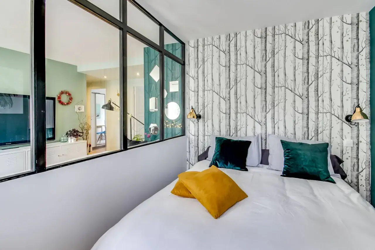 Id&eacute;e pour comment amenager une chambre en longueur : un lit douillet face &agrave; un mur v&eacute;g&eacute;tal stylis&eacute;, s&eacute;par&eacute; par une verri&egrave;re d'un salon lumineux.