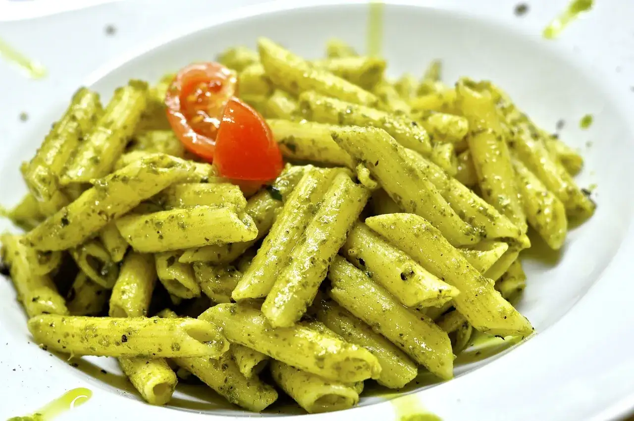 Un plato de pasta penne al pesto, con un trozo de tomate fresco encima.
