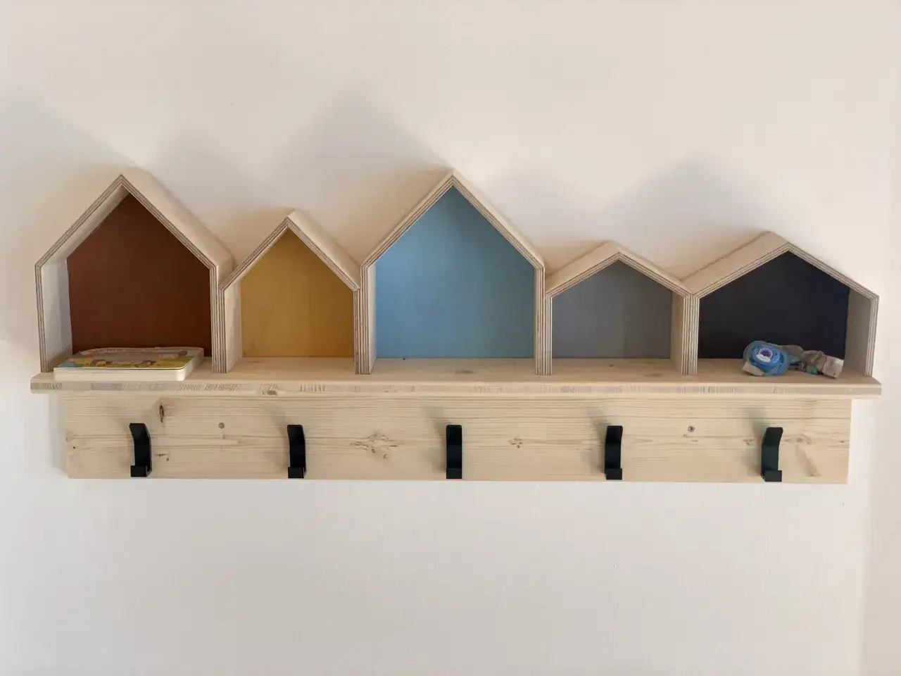 Selbstgebaute Kindergarderobe im Haus-Design mit Ablage und Haken. Ein Buch und ein Schnuller liegen auf der Ablage.
