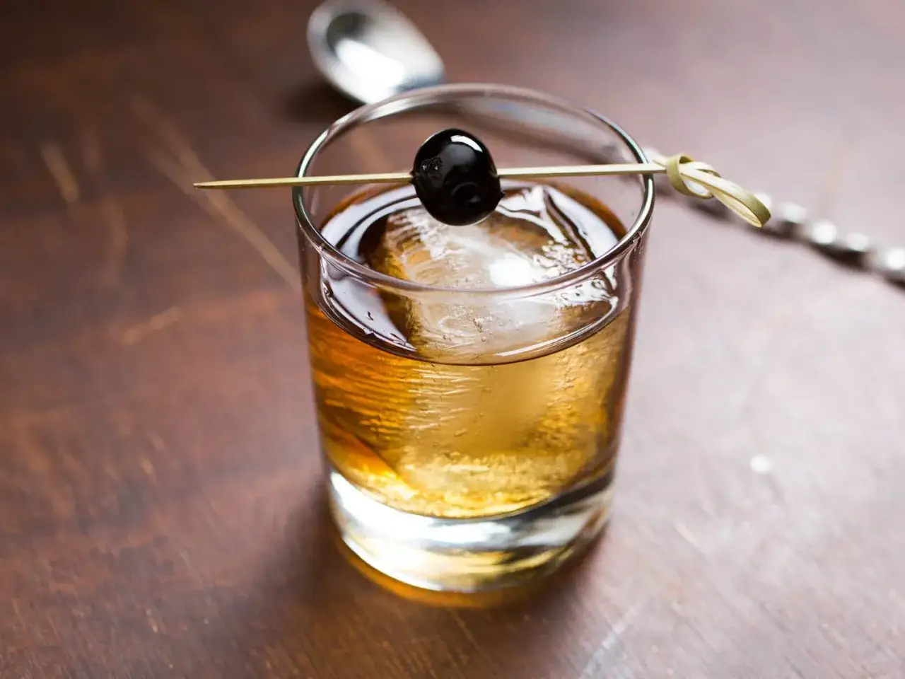 Klasyczny Sazerac z wiśnią na patyczku, lód i łyżka barmańska na drewnianym blacie.