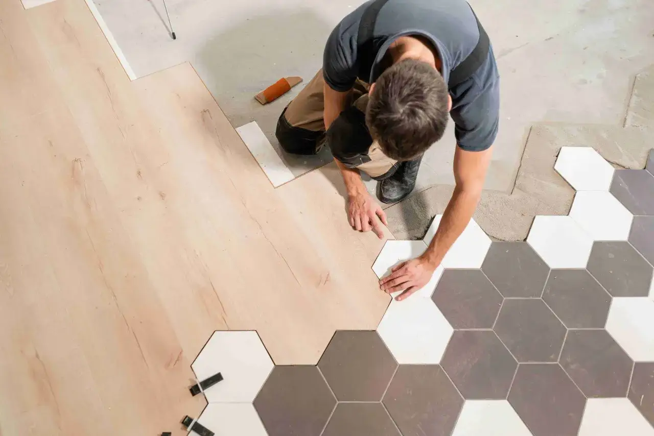 Un ouvrier pose du carrelage hexagonal blanc et gris &agrave; c&ocirc;t&eacute; d'un parquet en bois clair.