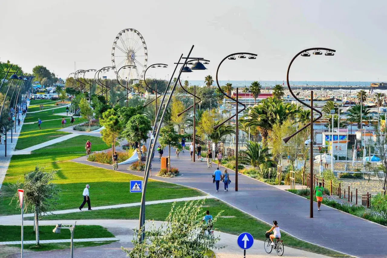 Passeggiata lungomare con gente che cammina e va in bici, tra verde e palme, con la ruota panoramica in lontananza, tipica di Rimini Riccione.
