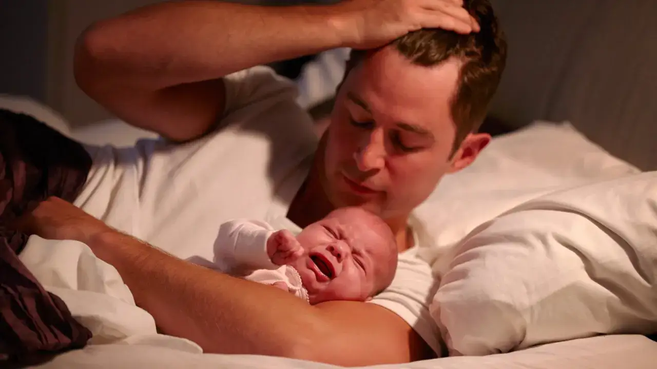 Ein Vater h&auml;lt sein weinendes Baby im Arm. Das Baby schluchzt im Schlaf, w&auml;hrend der Vater besorgt auf es herabblickt.