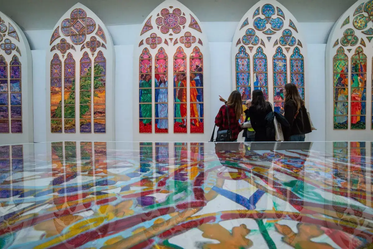 Trois personnes admirent des vitraux contemporains aux couleurs vives, leurs reflets se m&ecirc;lant &agrave; une &oelig;uvre au sol.