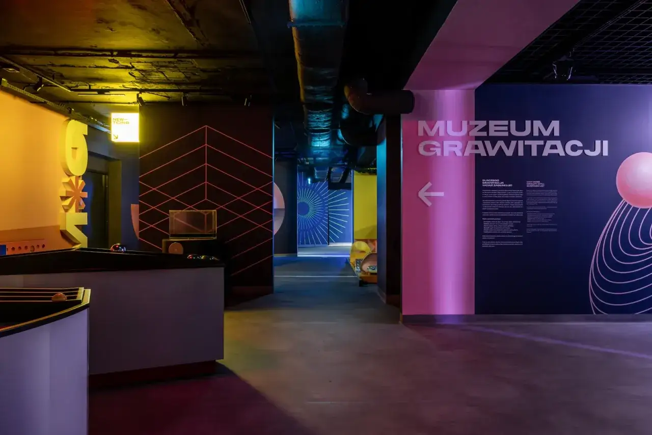 Kolorowe ściany i instalacje zapraszają do **Muzeum Grawitacji**. Widać tu tablice informacyjne i elementy interaktywne.