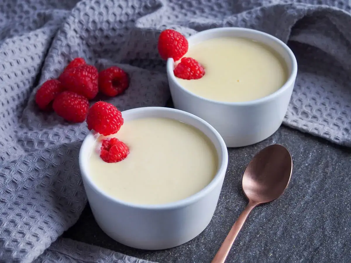 Dwa białe pucharki z kremowym deserem, ozdobione malinami. To jest właśnie custard, pyszny i delikatny.