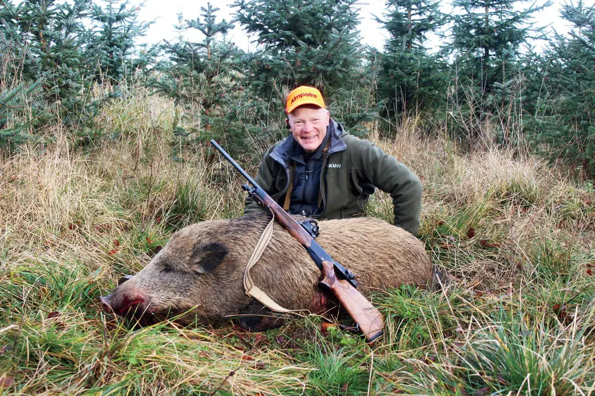Chasseur souriant avec un sanglier abattu, fusil à point rouge à portée de main. Une belle prise après une chasse réussie.