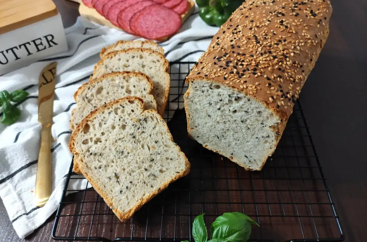 Chleb z sezamem, pokrojony w kromki, leży na kratce. Na wierzchu posypany białym i czarnym sezamem. Obok plasterki salami i zielona papryka.