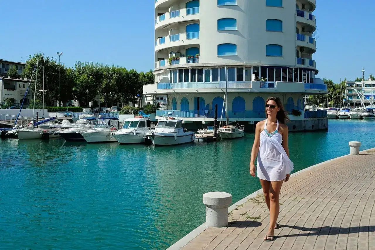Donna in bianco passeggia sul molo di un porto verde, con barche ormeggiate e un edificio bianco con balconi blu.
