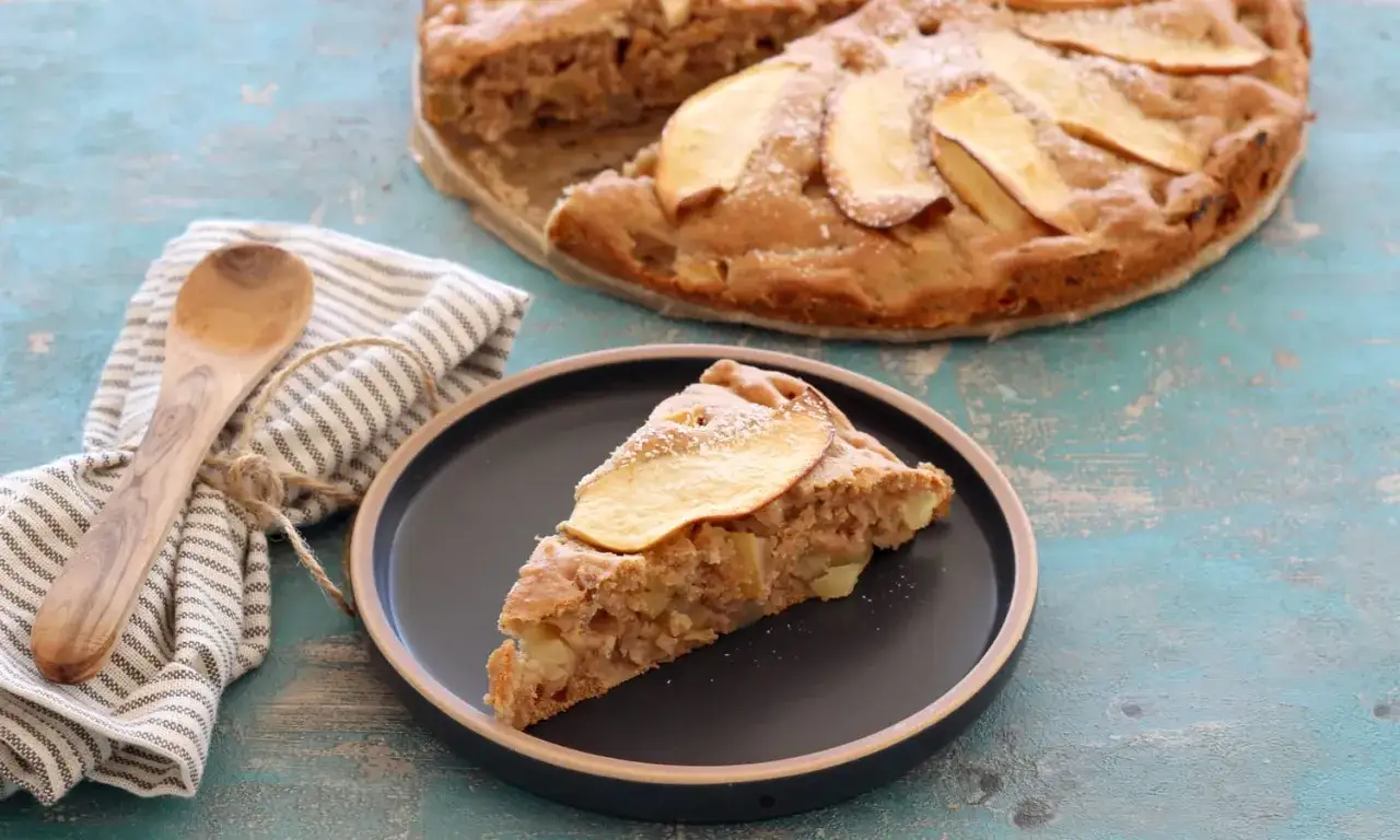Ein St&uuml;ck saftiger Apfelkuchen ohne Zucker, mit Apfelscheiben belegt und mit Puderzucker best&auml;ubt, auf einem Teller neben einem Holzl&ouml;ffel und einer Serviette.