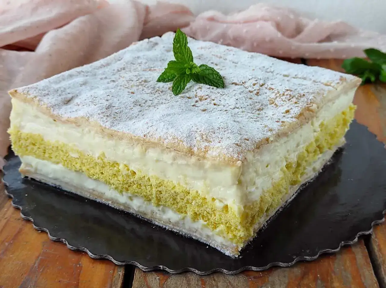 Una fetta di torta diplomatica, con pan di Spagna giallo, crema pasticcera e sfoglia, spolverata di zucchero a velo e guarnita con menta.