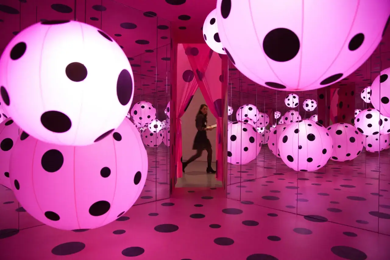 Salle rose et miroir, remplie de sph&egrave;res lumineuses &agrave; pois noirs, une femme traverse l'installation de Yayoi Kusama.