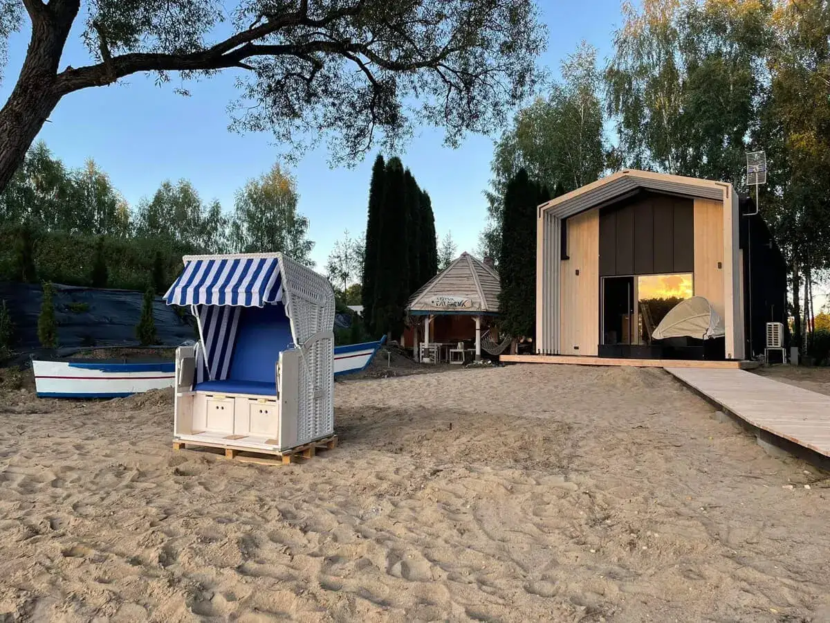 Piękny ośrodek wczasowy Mazury. Biały kosz plażowy z niebieskim daszkiem na piaszczystej plaży, obok łódź. W tle nowoczesny domek i altana.