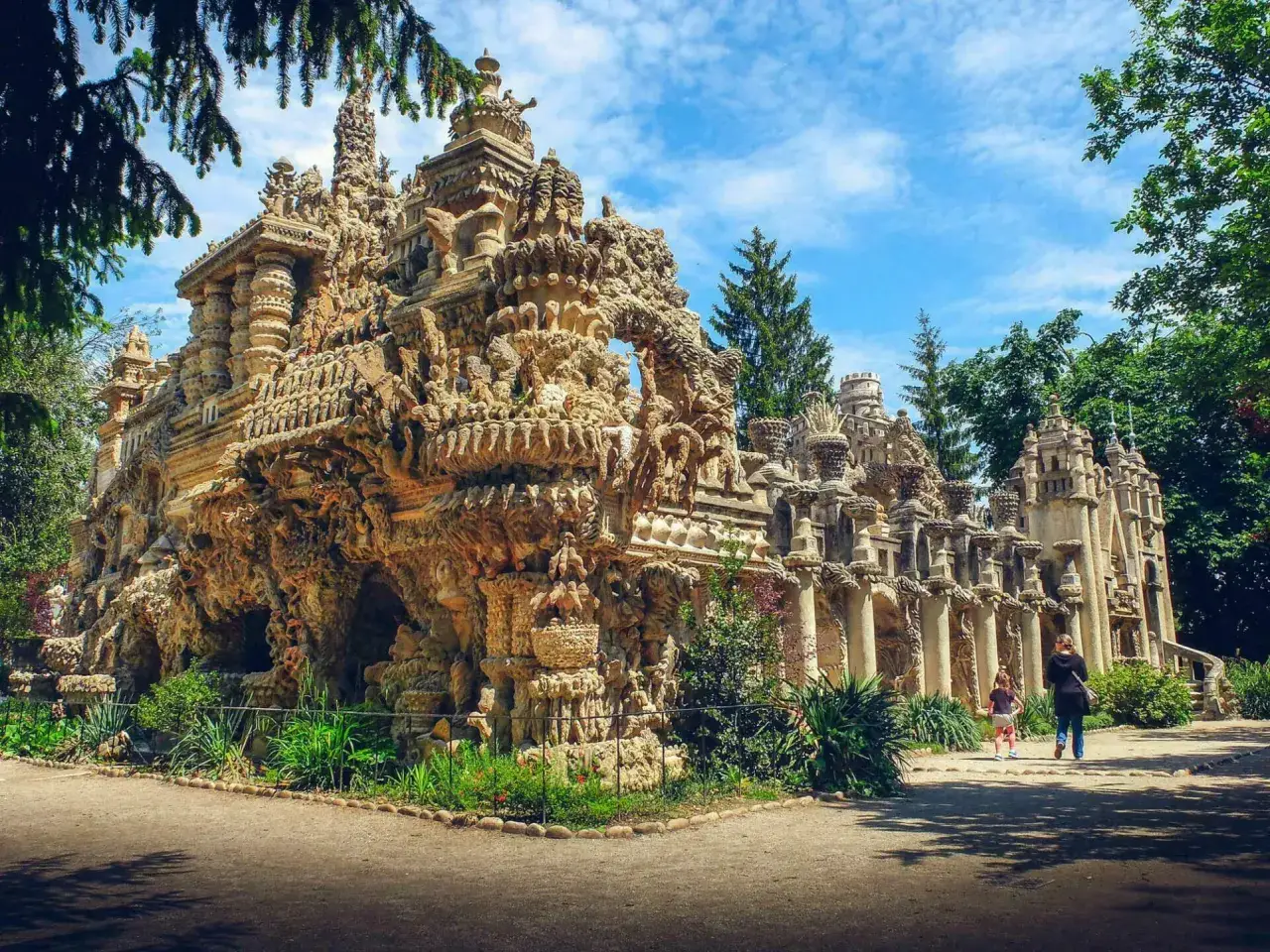 Une balade insolite dans un palais de pierre sculptée, où des détails fantastiques émergent sous un ciel bleu. Deux visiteurs découvrent ce lieu unique.