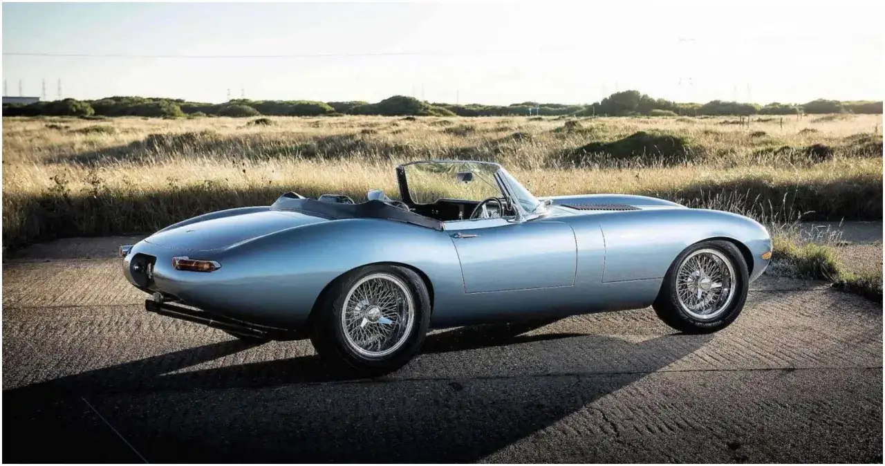 Błękitny kabriolet Jaguar E-Type, jeden z najpiękniejszych aut, na tle suchej trawy i nieba.