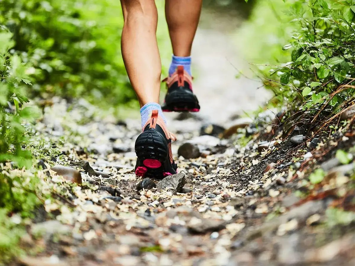 A person's legs in Merrell Nova 4 trail running shoes stride along a rocky, leaf-strewn path through lush green foliage.