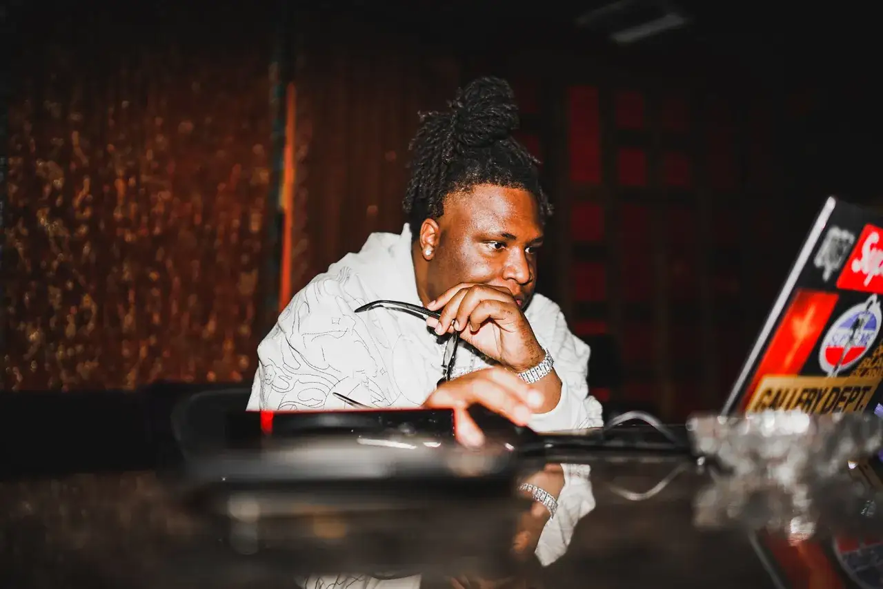 A young man with dreadlocks in a bun, wearing a white patterned shirt, looks thoughtfully at a laptop covered in stickers. He's holding glasses and appears to be working on new Tay Keith songs.