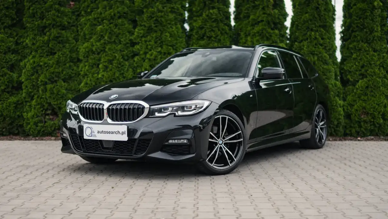 Eleganckie czarne BMW 320d kombi stoi przed żywopłotem.